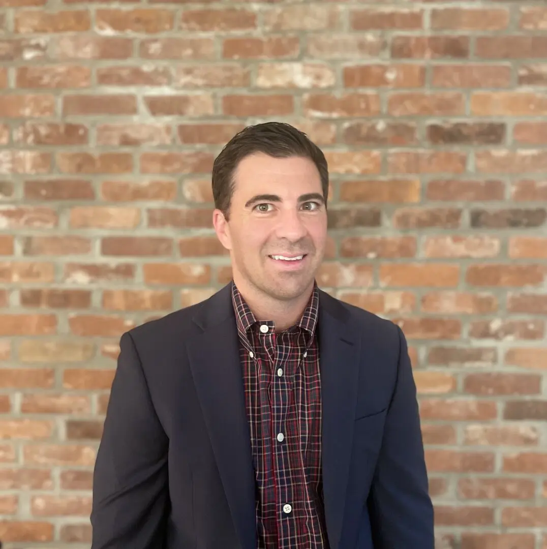 A man in a dark suit and plaid shirt stands smiling in front of a brick wall.