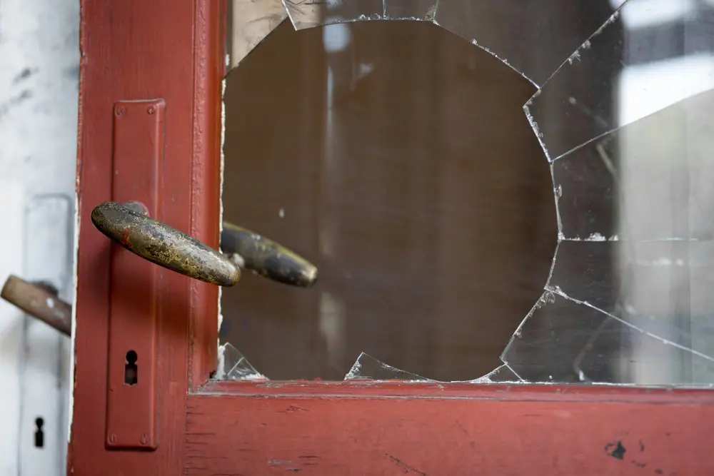 Red wooden door with a broken glass pane, showing a large hole. Two metal handles are visible, with a keyhole below the left handle.