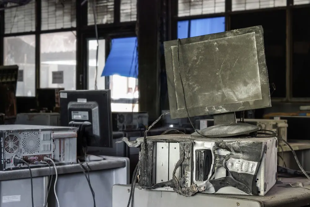 Damaged computer monitors and towers covered in dust and grime are piled in a dimly lit room.