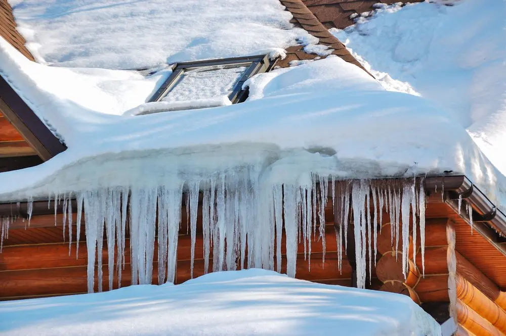 Icicles hanging from the snow-covered roof of a wooden building, with a skylight partially covered by snow.