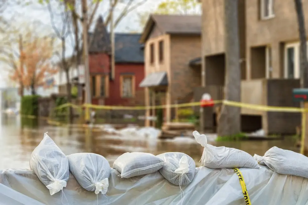 Sandbags lined up in front of houses during a flood, with streets partially submerged and caution tape in the background.