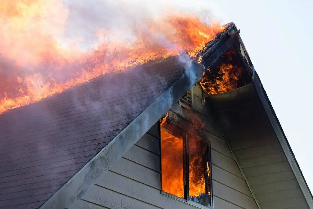 Fire engulfs the roof and window of a house, with flames and smoke visible against the sky.