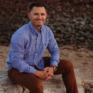 Man in a plaid shirt and brown pants sits on a rock outdoors, smiling at the camera. Rocky terrain is visible in the background.