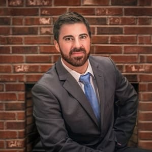 A man in a suit with a blue tie sits in front of a brick wall.