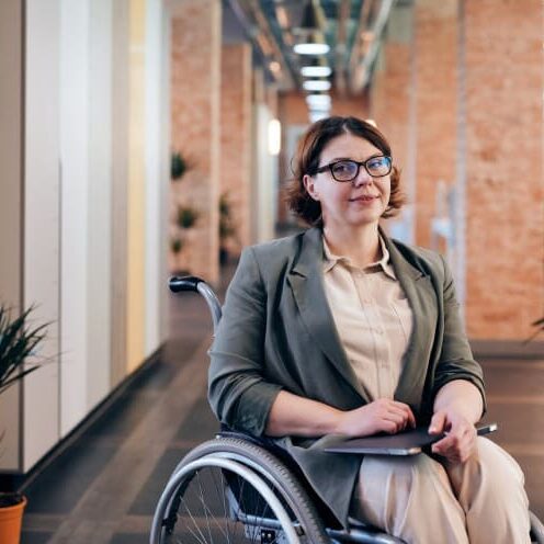 A person in a wheelchair holding a tablet in an office hallway with brick walls and plants.